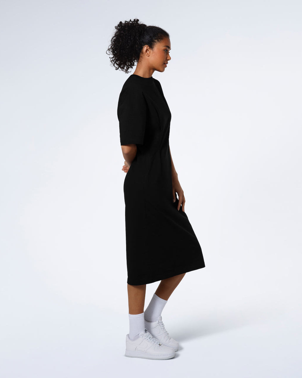A women standing side on modelling a plain black, organic cotton midi t shirt dress.