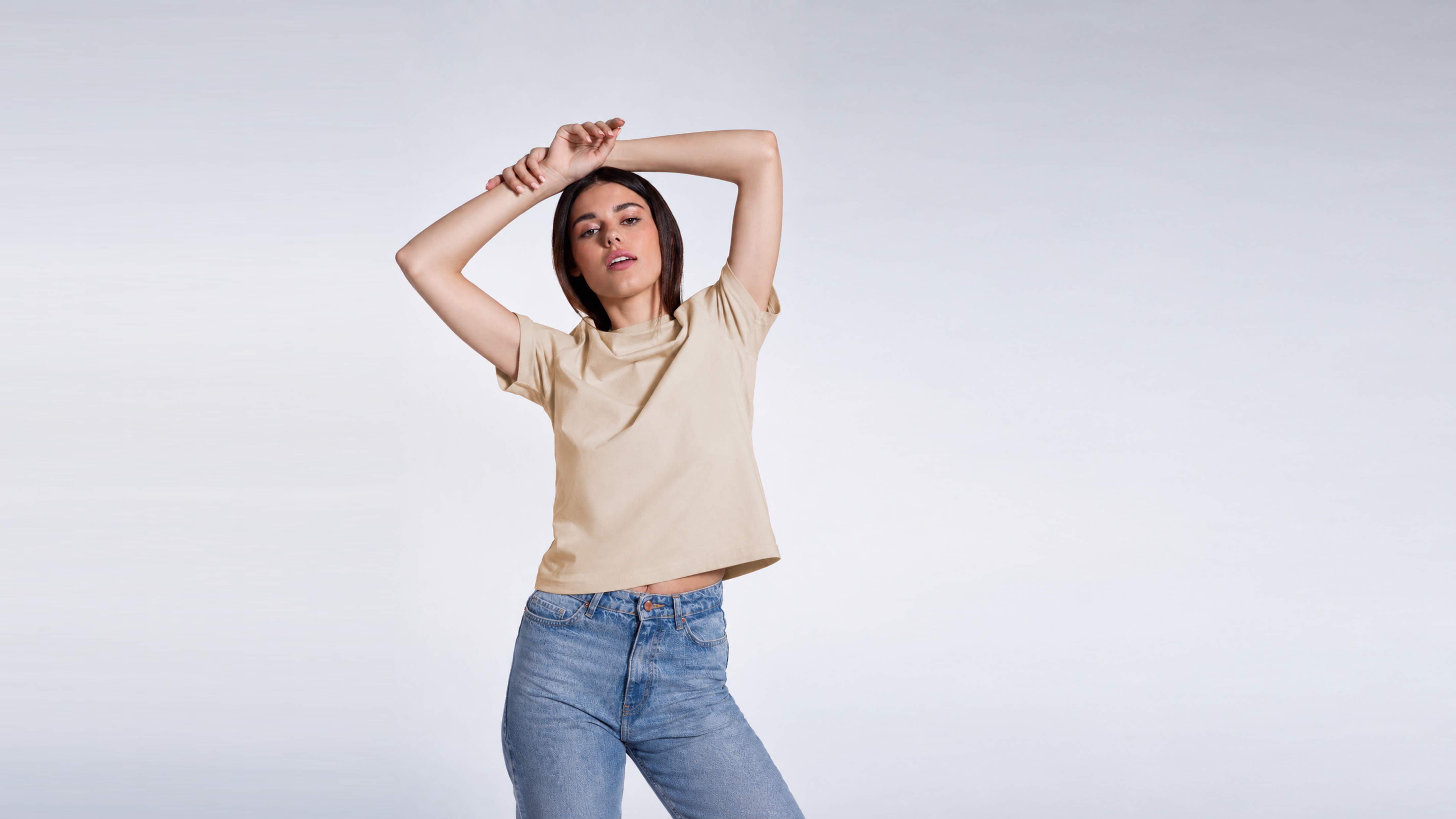 A woman looking into the camera with her arms over her head, modelling a plain beige, organic cotton t shirt.