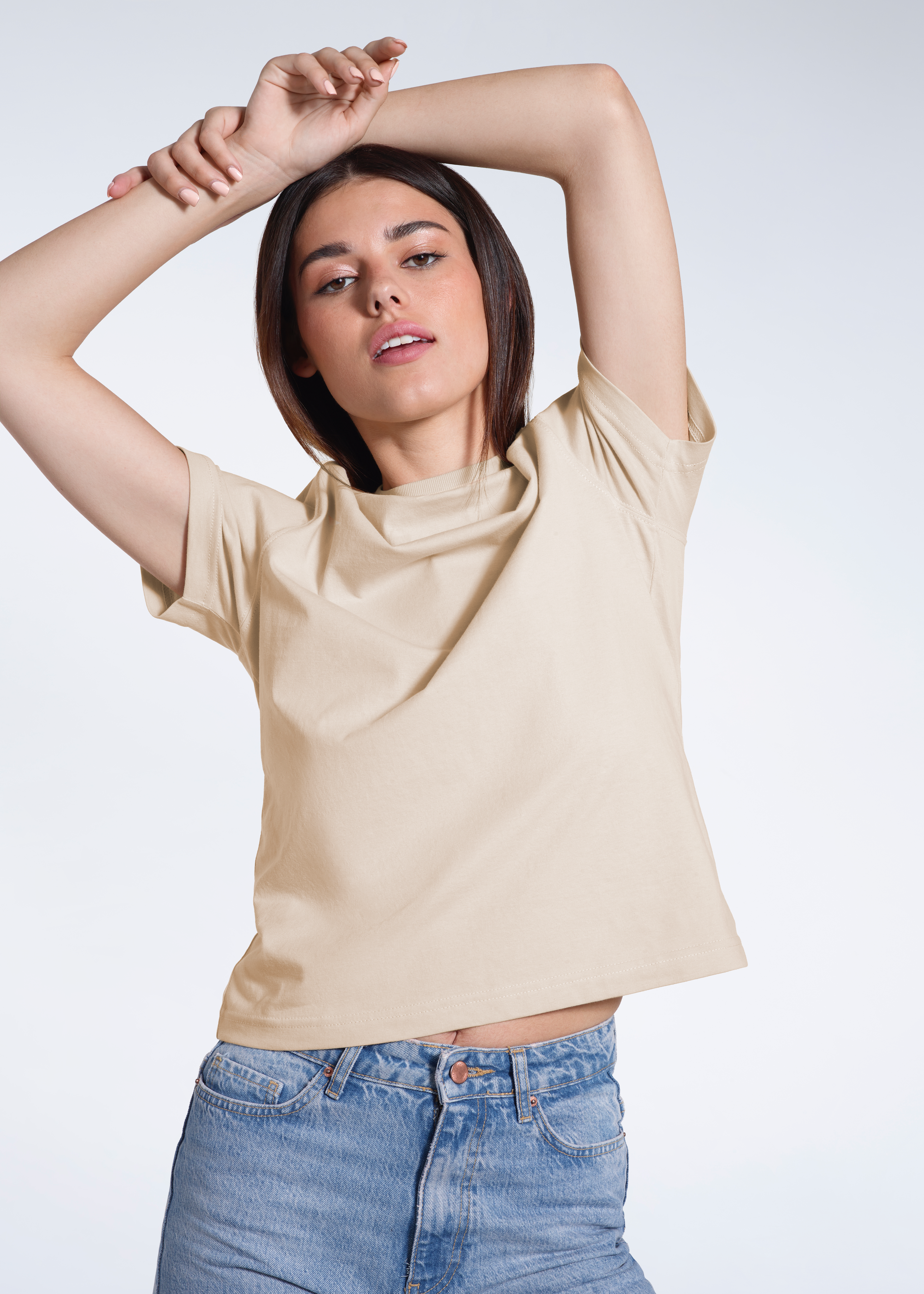 A woman looking into the camera with her arms over her head, modelling a plain beige, organic cotton t shirt.