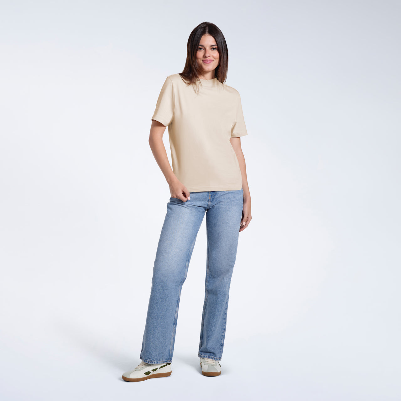 A full body photo of a woman looking into the camera smiling, modelling a plain beige short sleeve organic cotton t shirt.