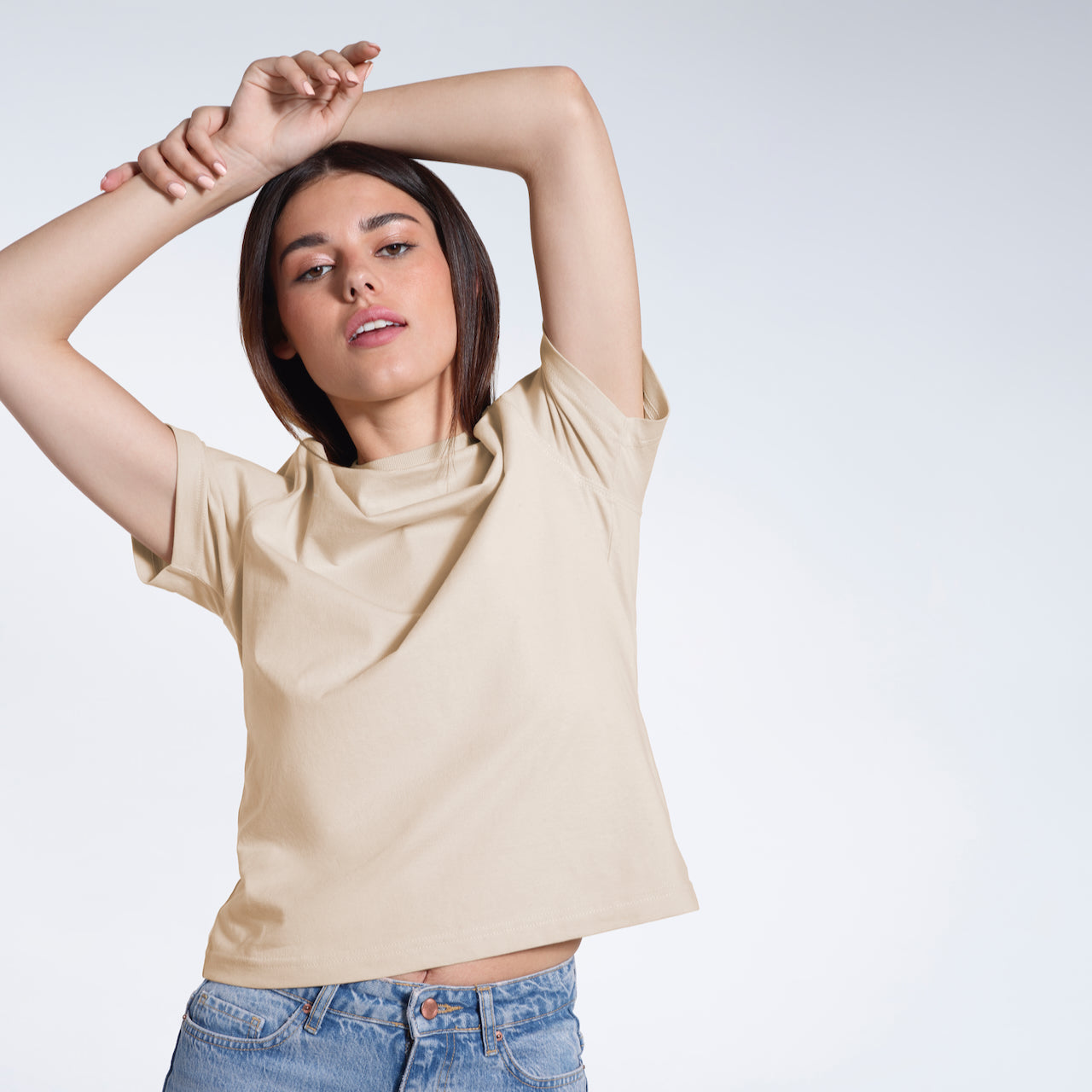 A woman with her hands above her head looking into the camera, modelling a plain beige short sleeve organic cotton t shirt.