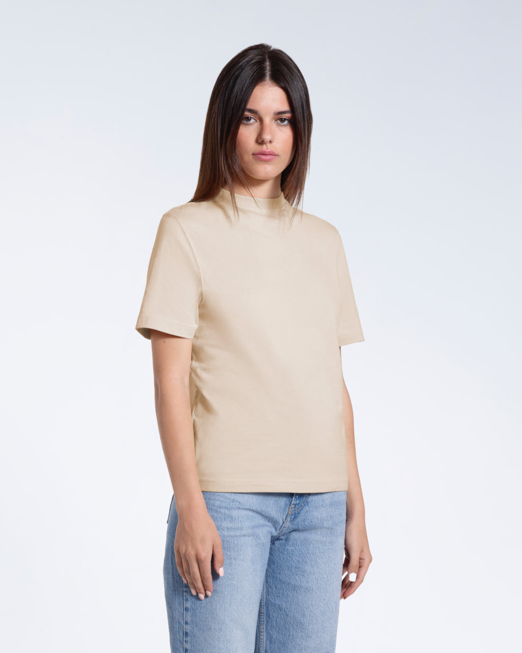 A woman looking into the camera with her arms by her sides, modelling a plain beige organic cotton t shirt.