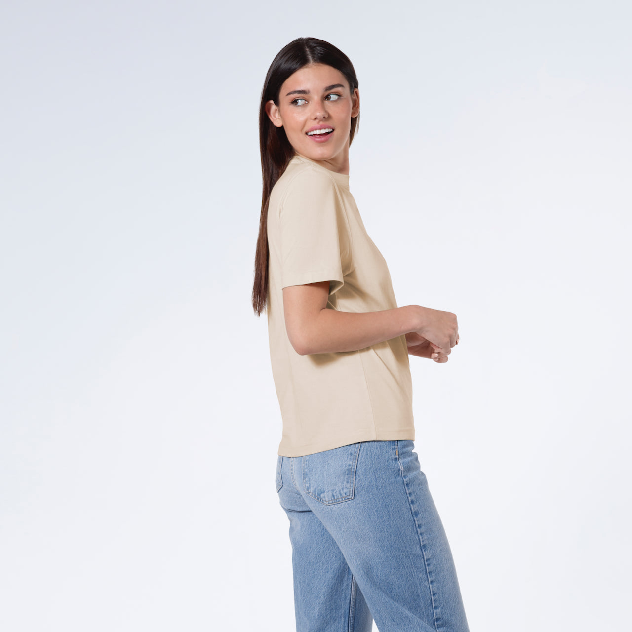 A woman facing side on and looking behind her smiling, modelling a plain beige short sleeve organic cotton t shirt.
