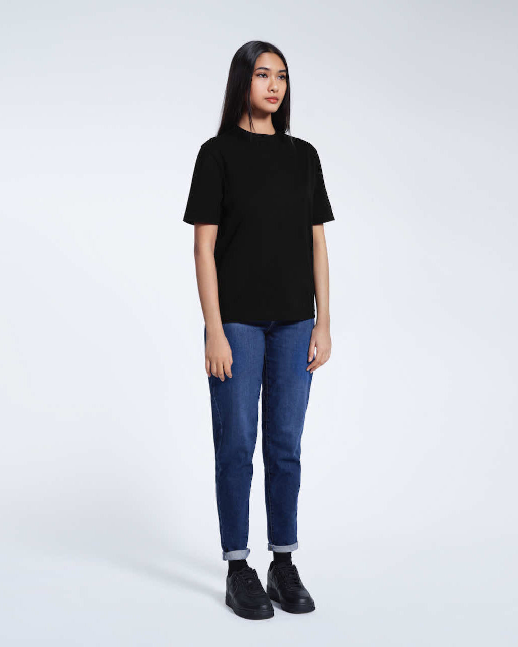 A full body photo of a woman with her arms at her sides, modelling a plain black short sleeve organic cotton t shirt.
