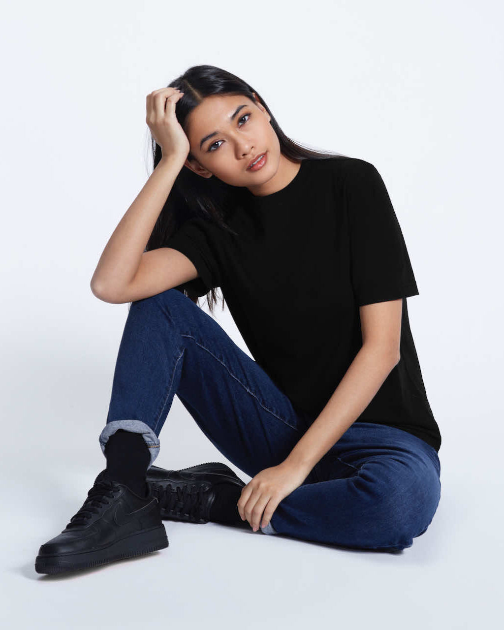 A woman sitting on the floor looking into the camera, modelling a plain black short sleeve organic cotton t shirt.