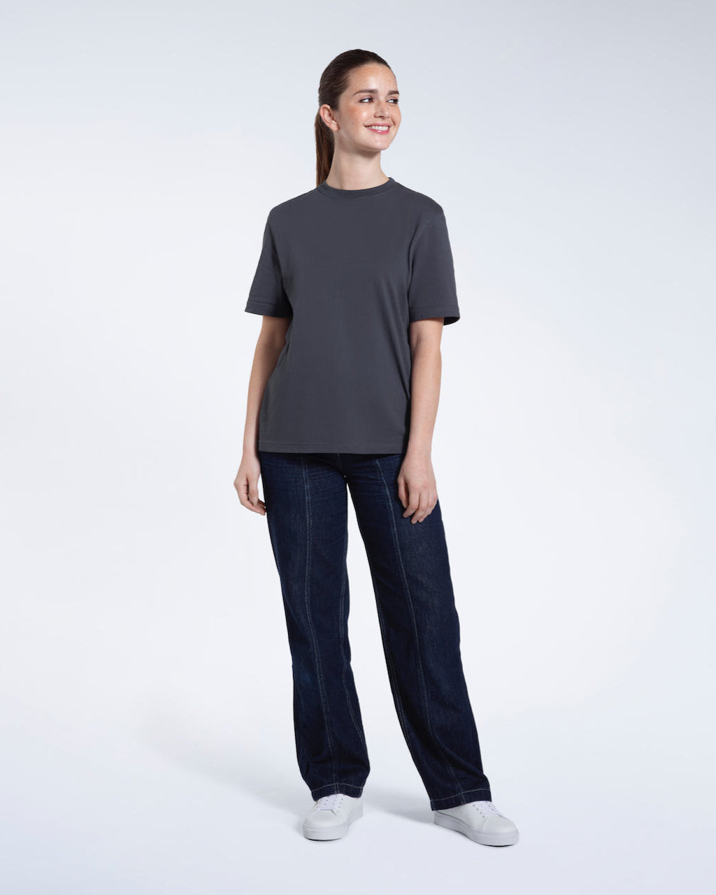 A full body photo of a woman smiling with her arms by her sides, modelling a plain dark grey short sleeve organic cotton t shirt.