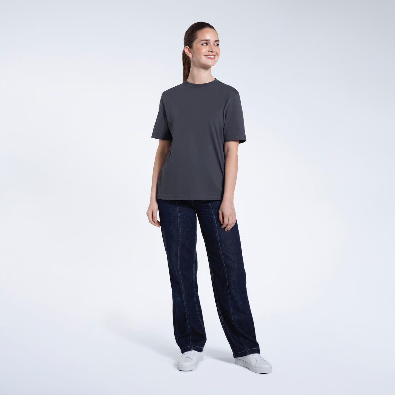 A full body photo of a woman smiling with her arms by her sides, modelling a plain dark grey short sleeve organic cotton t shirt.