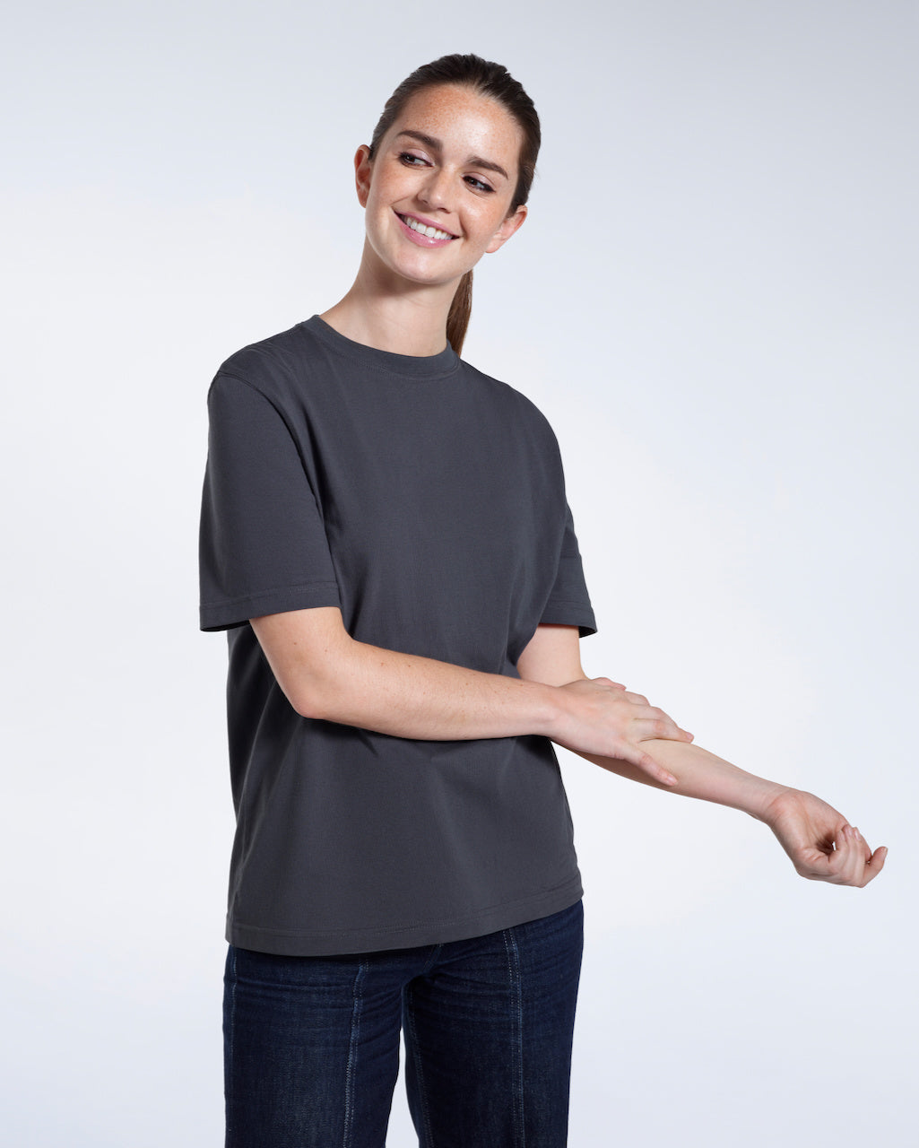 A woman smiling touching her arm, modelling a plain dark grey short sleeve organic cotton t shirt.