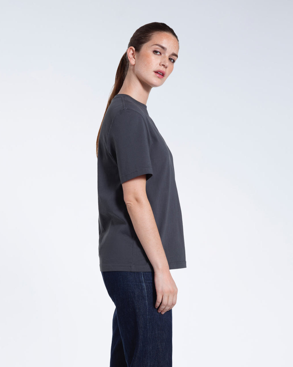 A woman facing side on and looking at the camera, modelling a plain dark grey short sleeve organic cotton t shirt.
