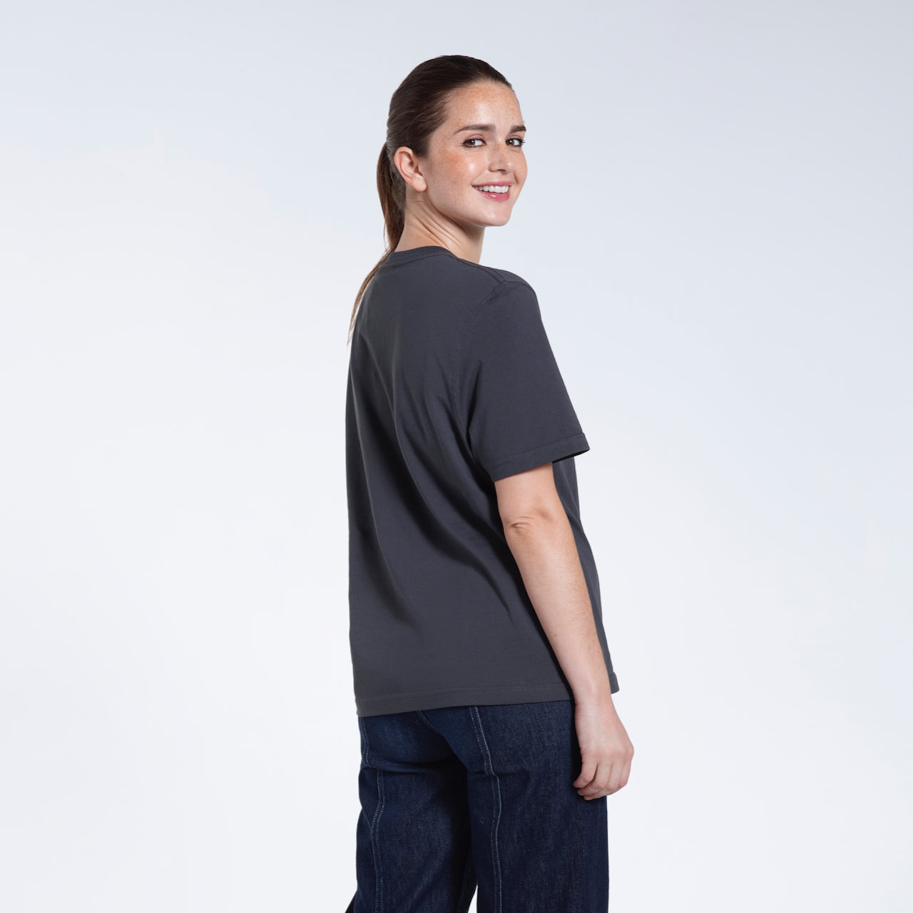 A woman turned towards the camera smiling, modelling a plain dark grey short sleeve organic cotton t shirt.