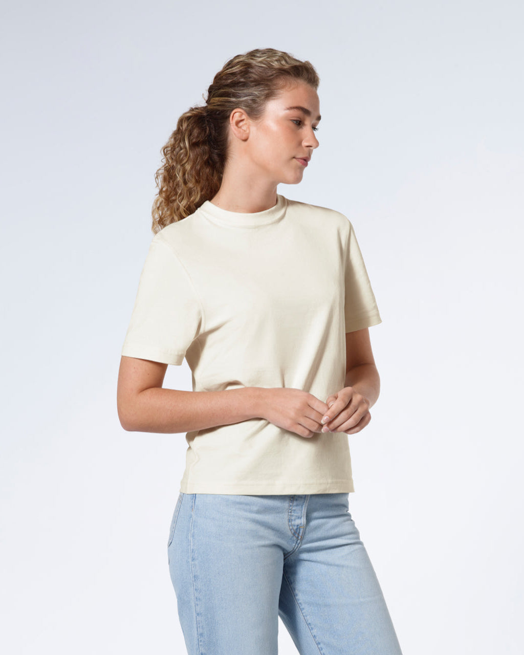 A woman with her hands together looking away from the camera, modelling a plain ecru short sleeve organic cotton t shirt.