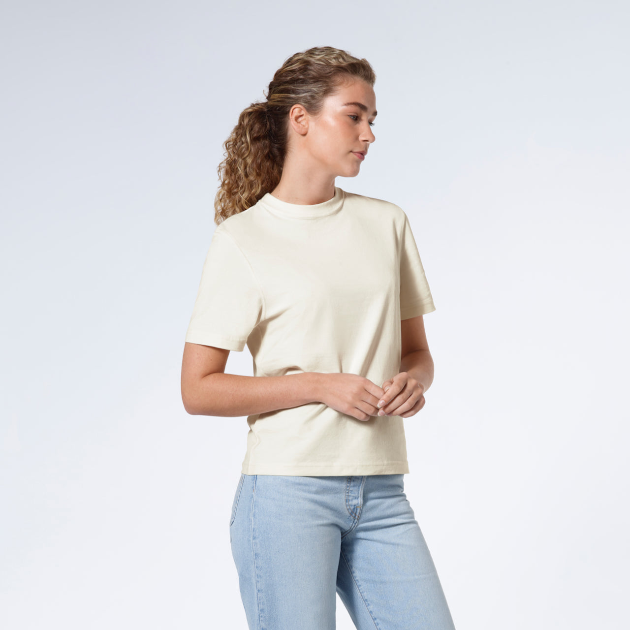 A woman with her hands together looking away from the camera, modelling a plain ecru short sleeve organic cotton t shirt.
