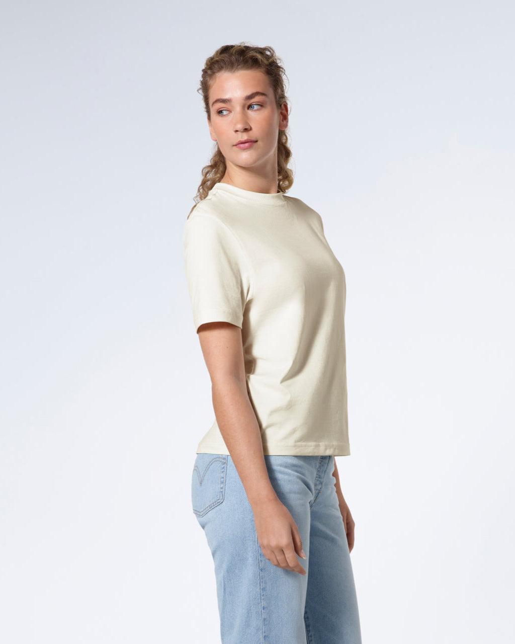 A woman facing side on looking back, modelling a plain ecru short sleeve organic cotton t shirt.