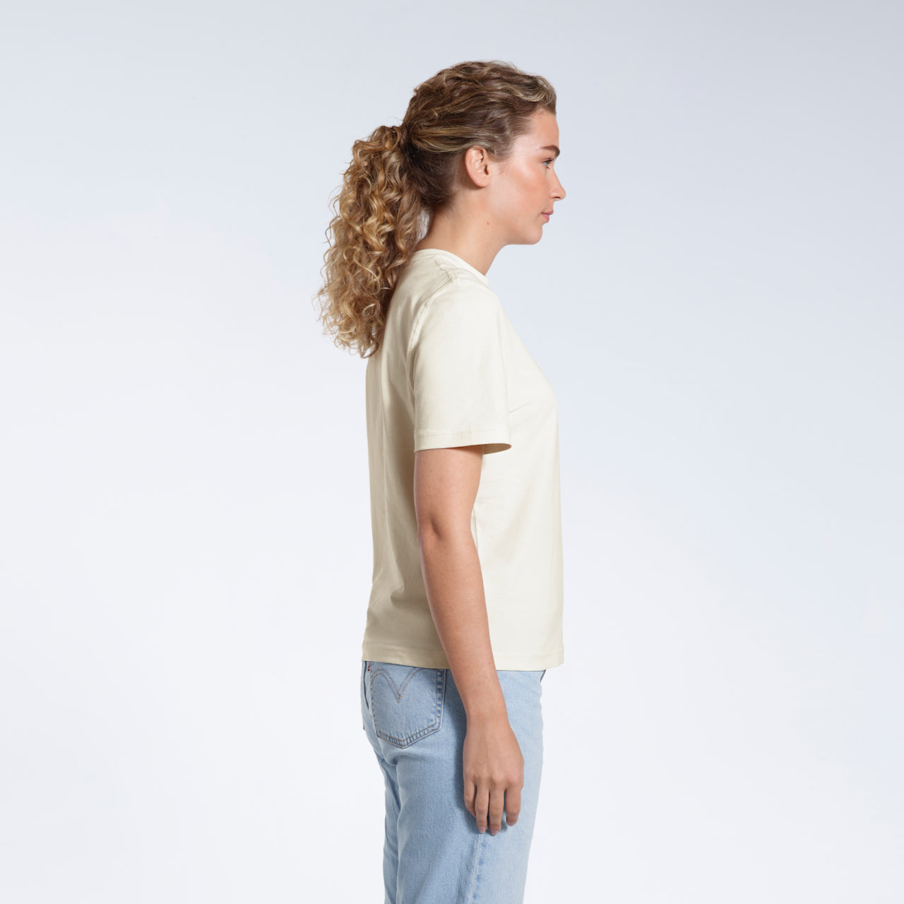 A woman facing side on, modelling a plain ecru short sleeve organic cotton t shirt.