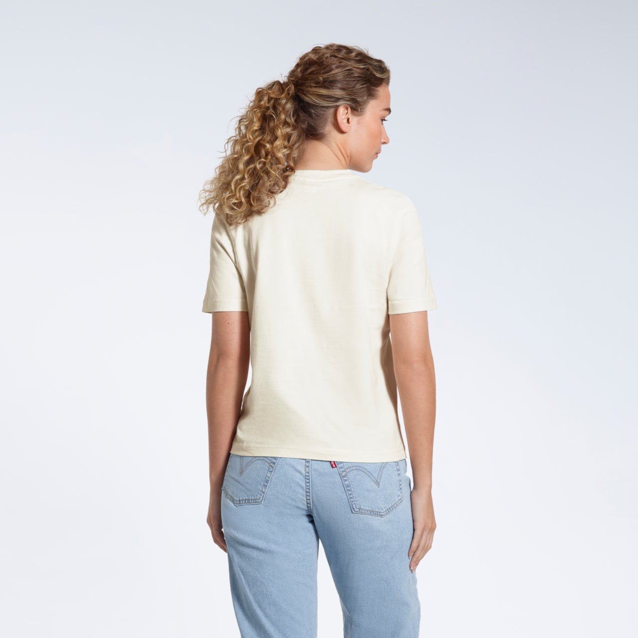 A woman with her back to the camera, modelling a plain ecru short sleeve organic cotton t shirt.