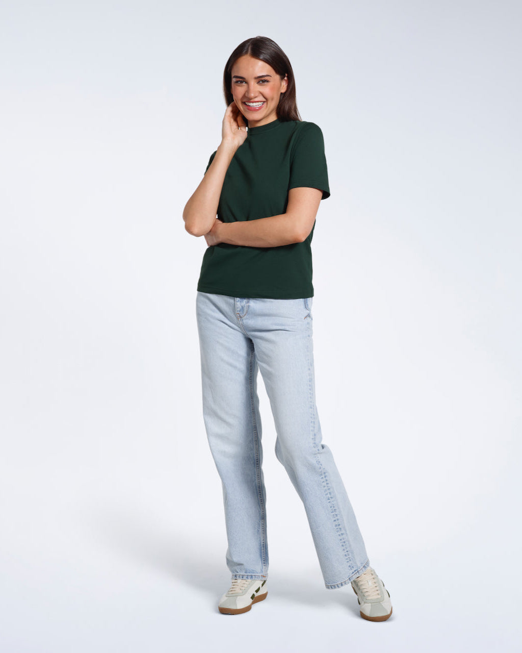 A full body photo of a woman touching her face smiling, modelling a plain green short sleeve organic cotton t shirt.