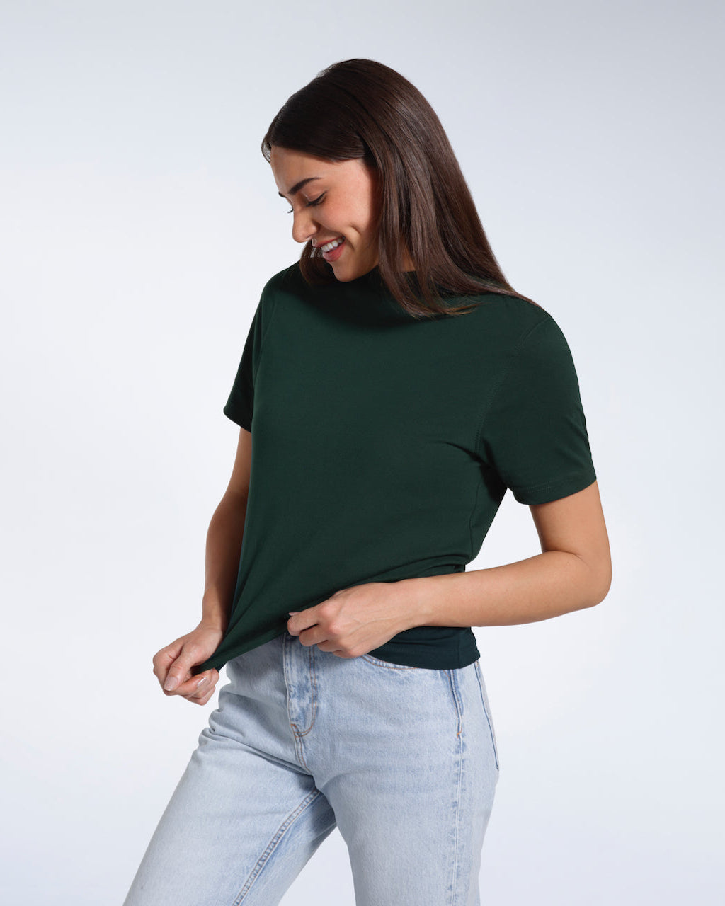 A woman looking down smiling and touching the bottom of her plain green short sleeve organic cotton t shirt.