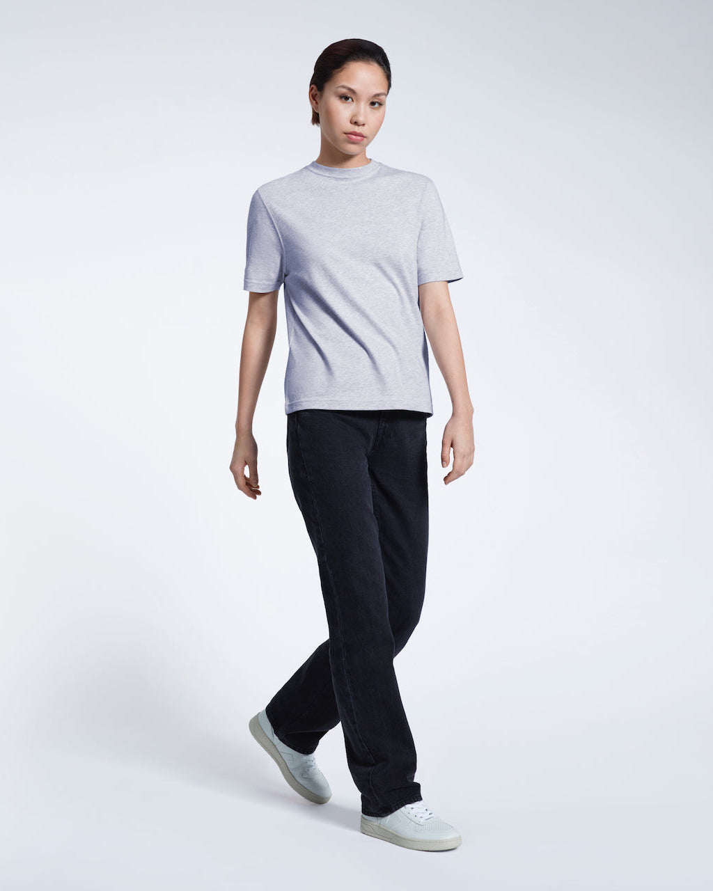 A full body photo of a woman walking looking into the camera, modelling a plain grey short sleeve organic cotton t shirt.