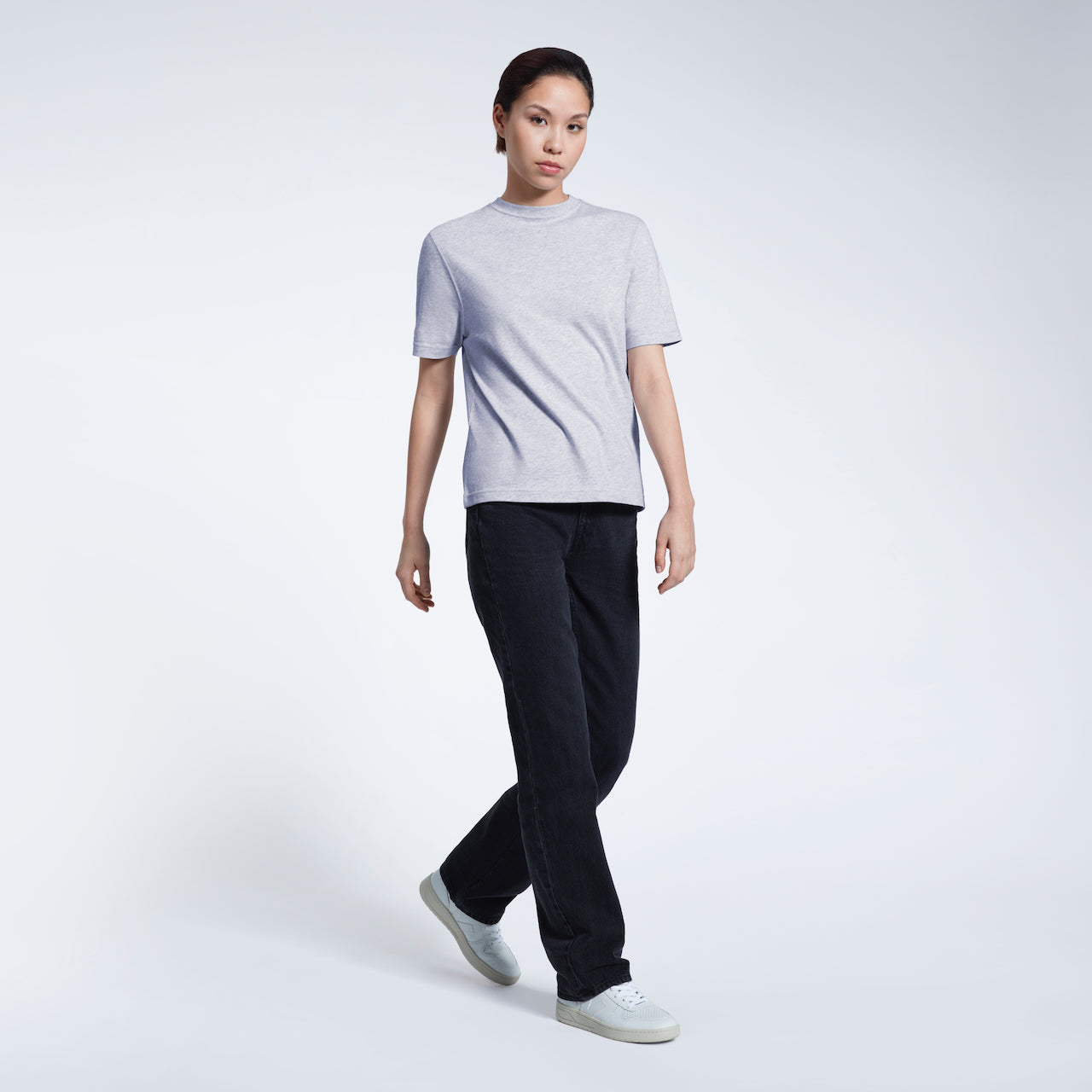 A full body photo of a woman walking looking into the camera, modelling a plain grey short sleeve organic cotton t shirt.