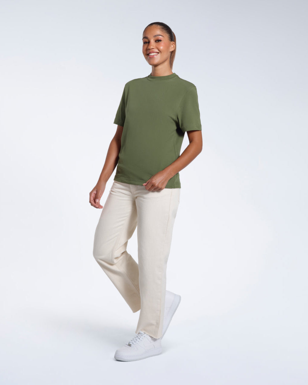 A full body photo of a woman walking and smiling, modelling a plain khaki green short sleeve organic cotton t shirt.