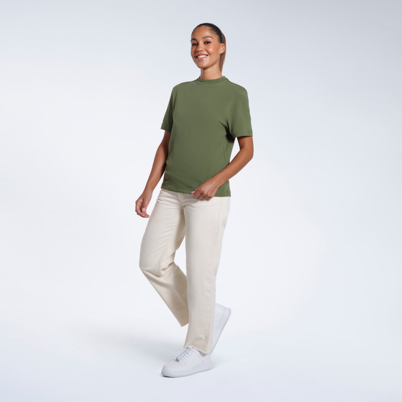 A full body photo of a woman walking and smiling, modelling a plain khaki green short sleeve organic cotton t shirt.