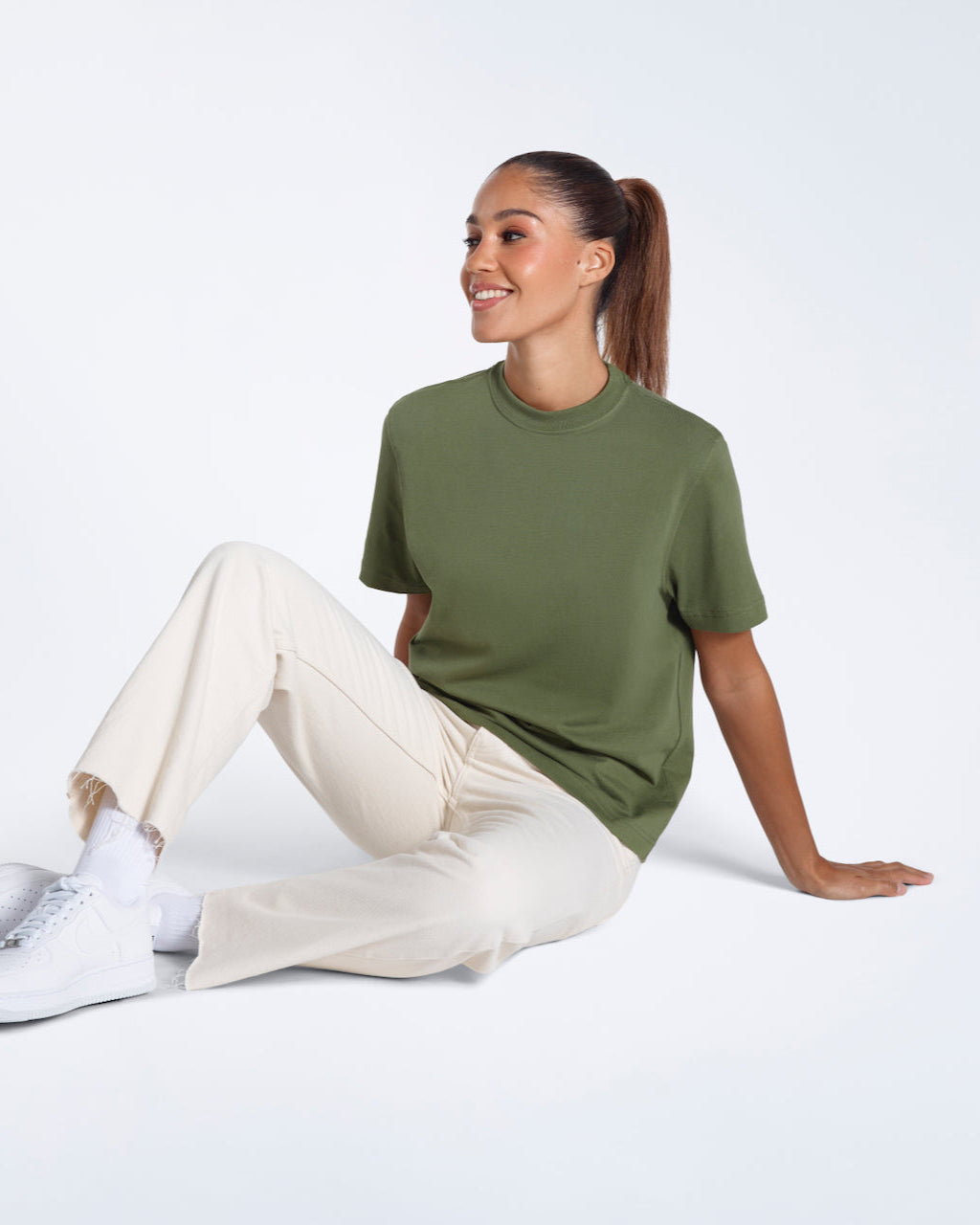 A woman sitting on the floor smiling, modelling a plain khaki green short sleeve organic cotton t shirt.