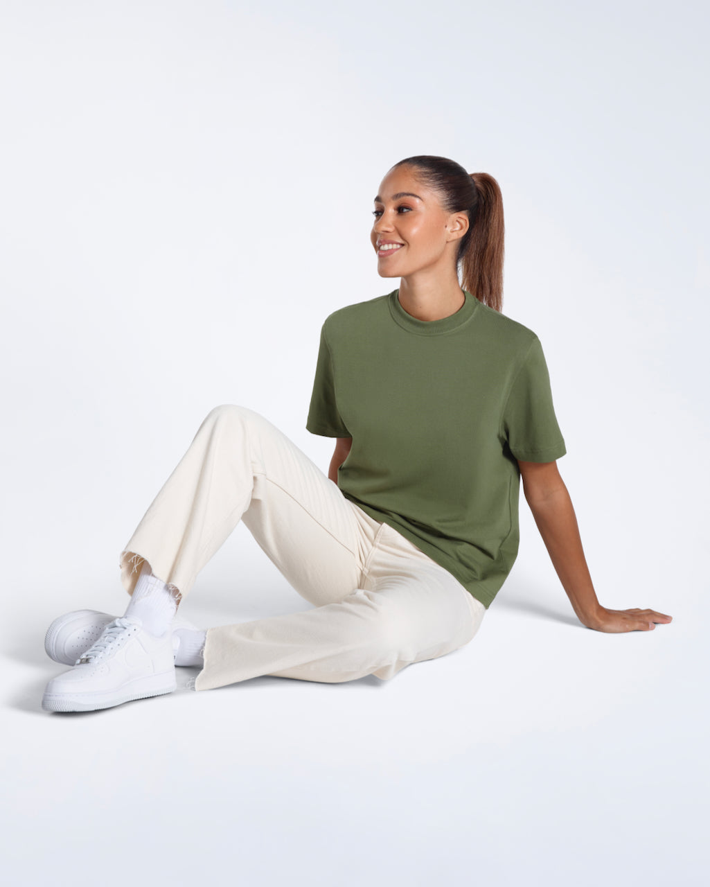 A woman sitting on the floor smiling, modelling a plain khaki green short sleeve organic cotton t shirt.