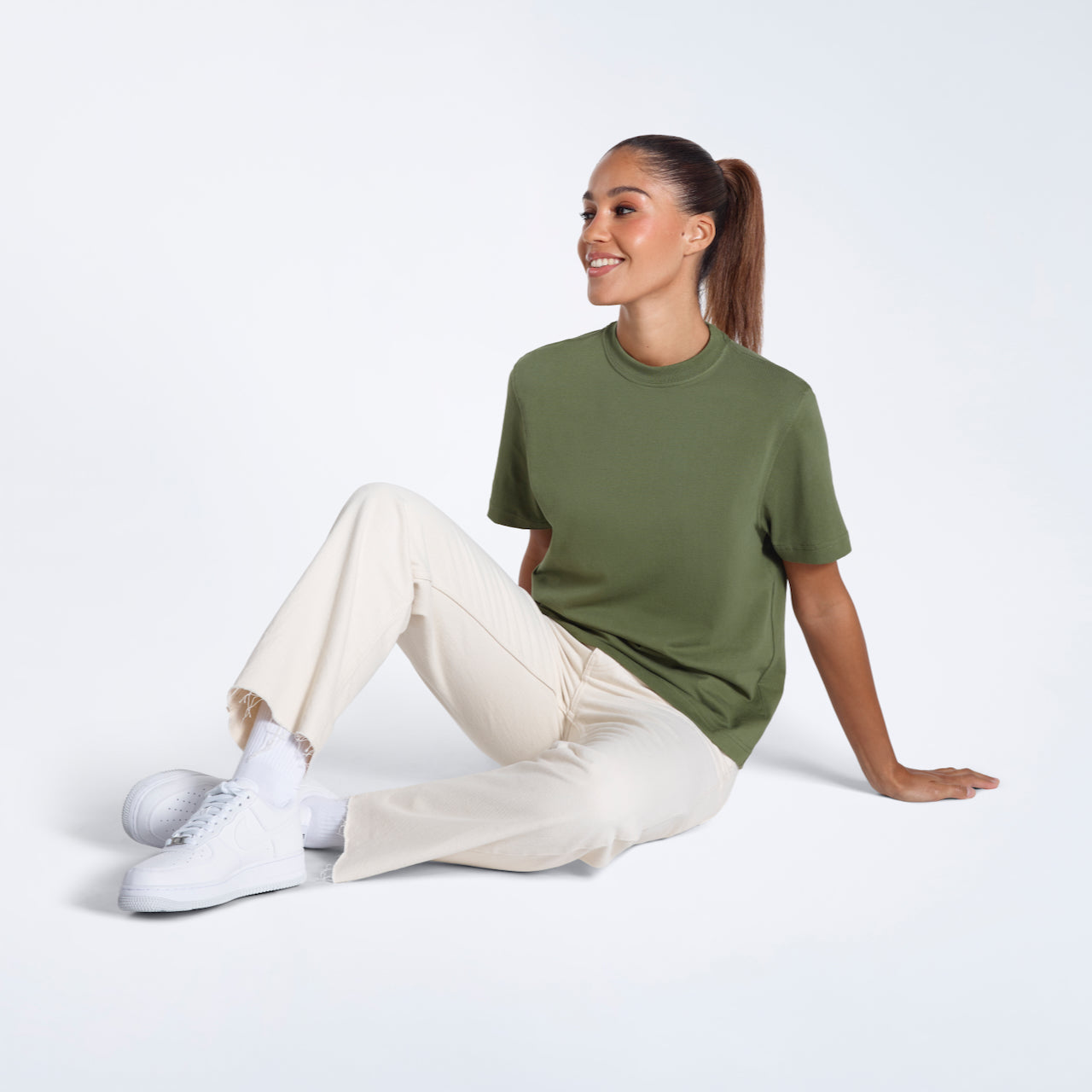 A woman sitting on the floor smiling, modelling a plain khaki green short sleeve organic cotton t shirt.