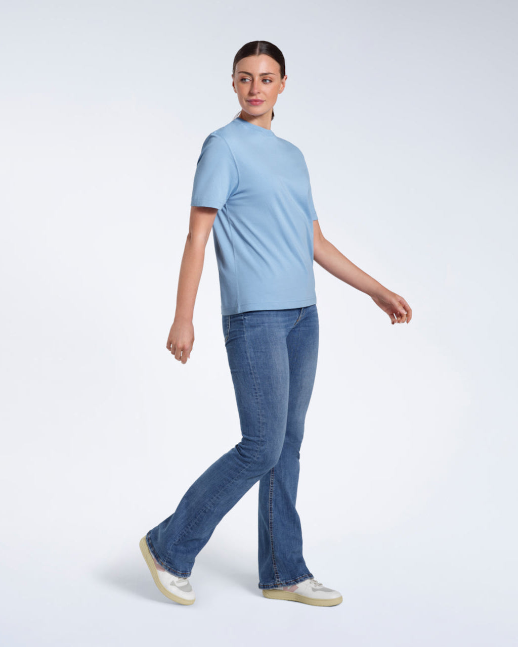 A full body photo of a woman walking looking to her side, modelling a plain blue short sleeve organic cotton t shirt.