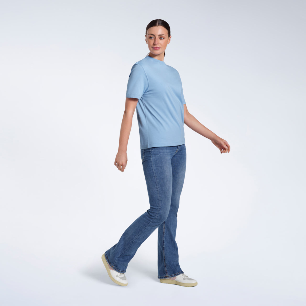 A full body photo of a woman walking looking to her side, modelling a plain blue short sleeve organic cotton t shirt.