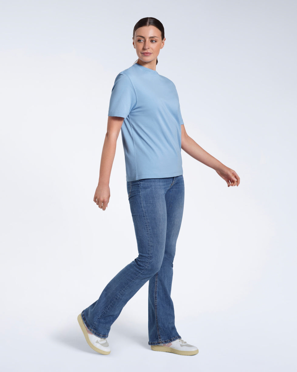 A full body photo of a woman walking looking to her side, modelling a plain blue short sleeve organic cotton t shirt.
