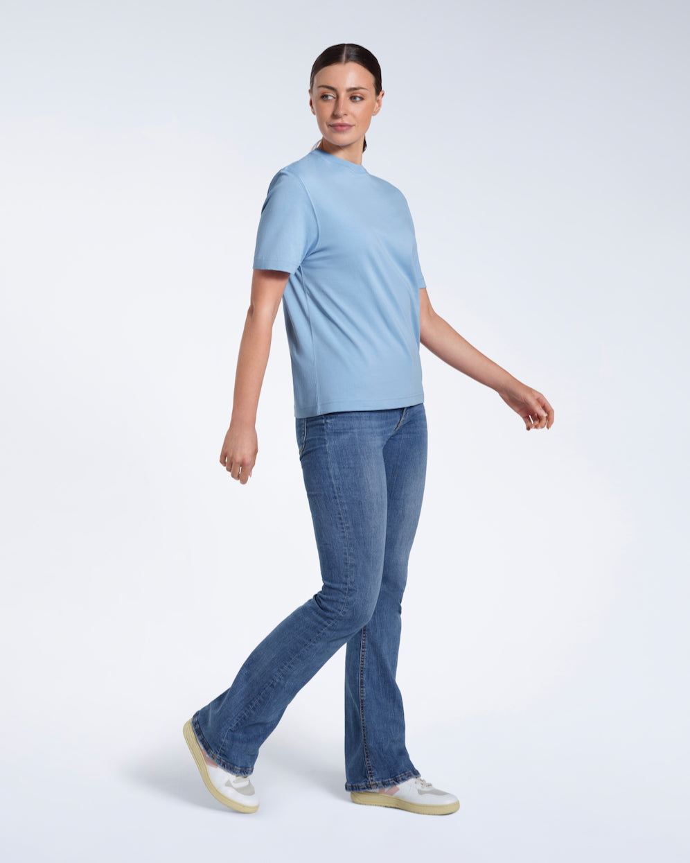 A full body photo of a woman walking looking to her side, modelling a plain blue short sleeve organic cotton t shirt.