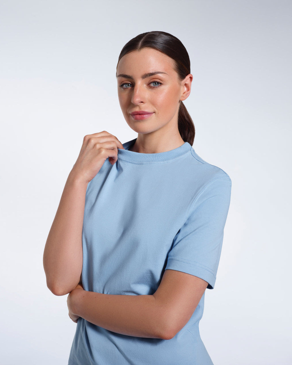 A woman looking into the camera smiling and touching her collar, modelling a plain blue short sleeve organic cotton t shirt.