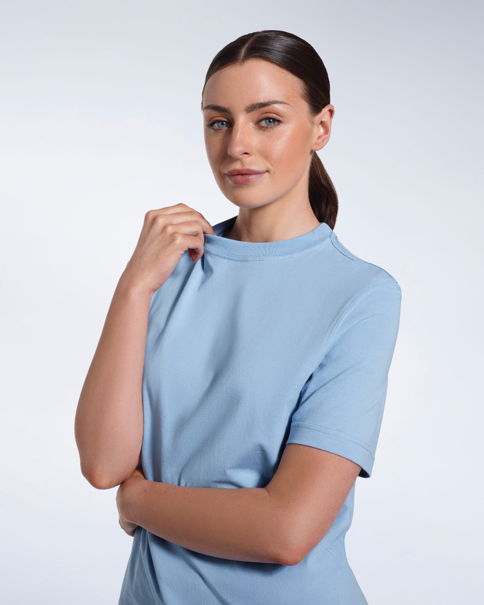 A woman looking into the camera smiling and touching her collar, modelling a plain blue short sleeve organic cotton t shirt.