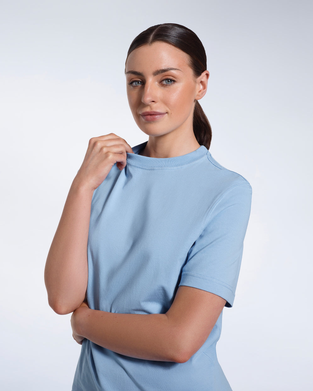 A woman looking into the camera smiling and touching her collar, modelling a plain blue short sleeve organic cotton t shirt.