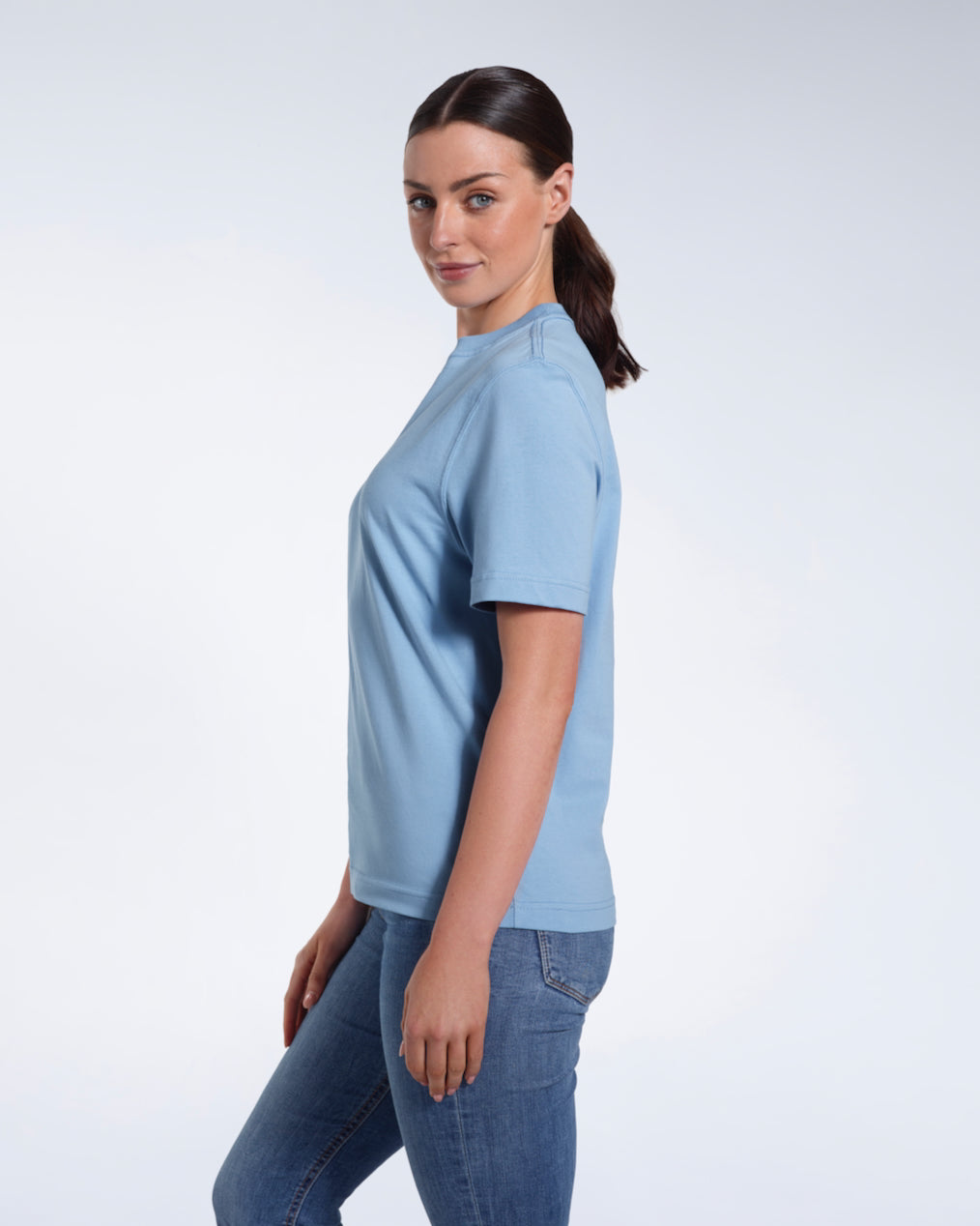 A woman facing side on and looking at the camera, modelling a plain blue short sleeve organic cotton t shirt.