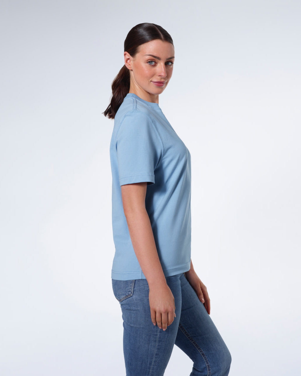 A woman facing side on and looking at the camera, modelling a plain blue short sleeve organic cotton t shirt.