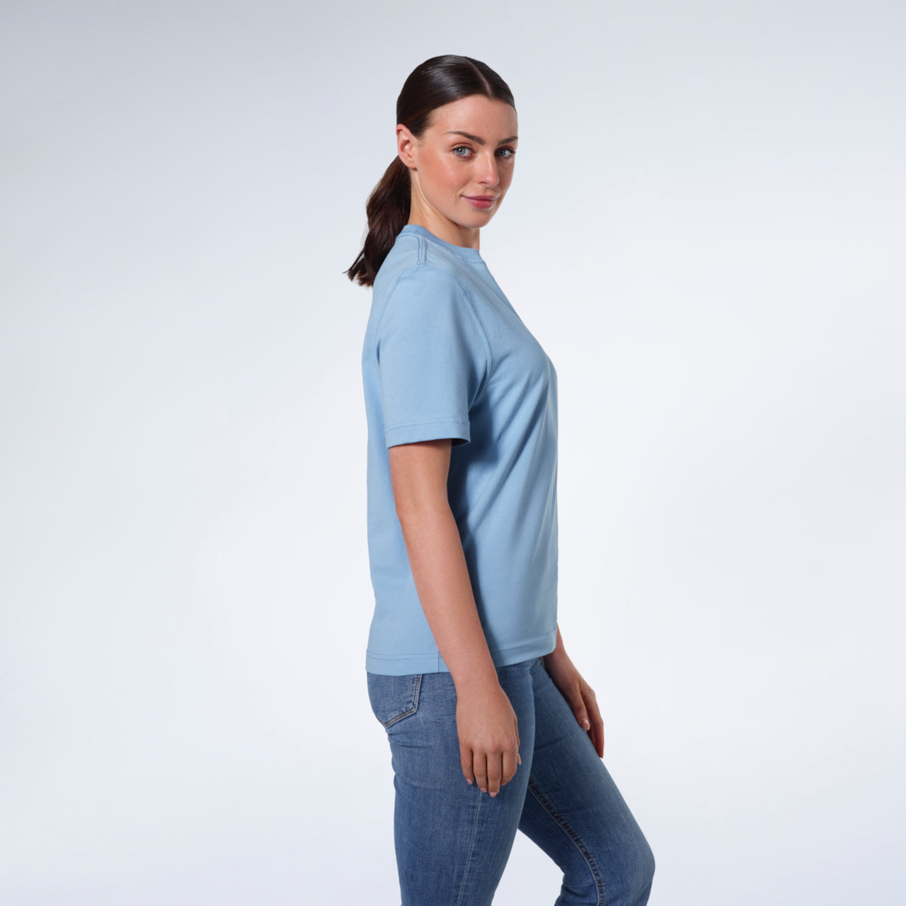 A woman facing side on and looking at the camera, modelling a plain blue short sleeve organic cotton t shirt.