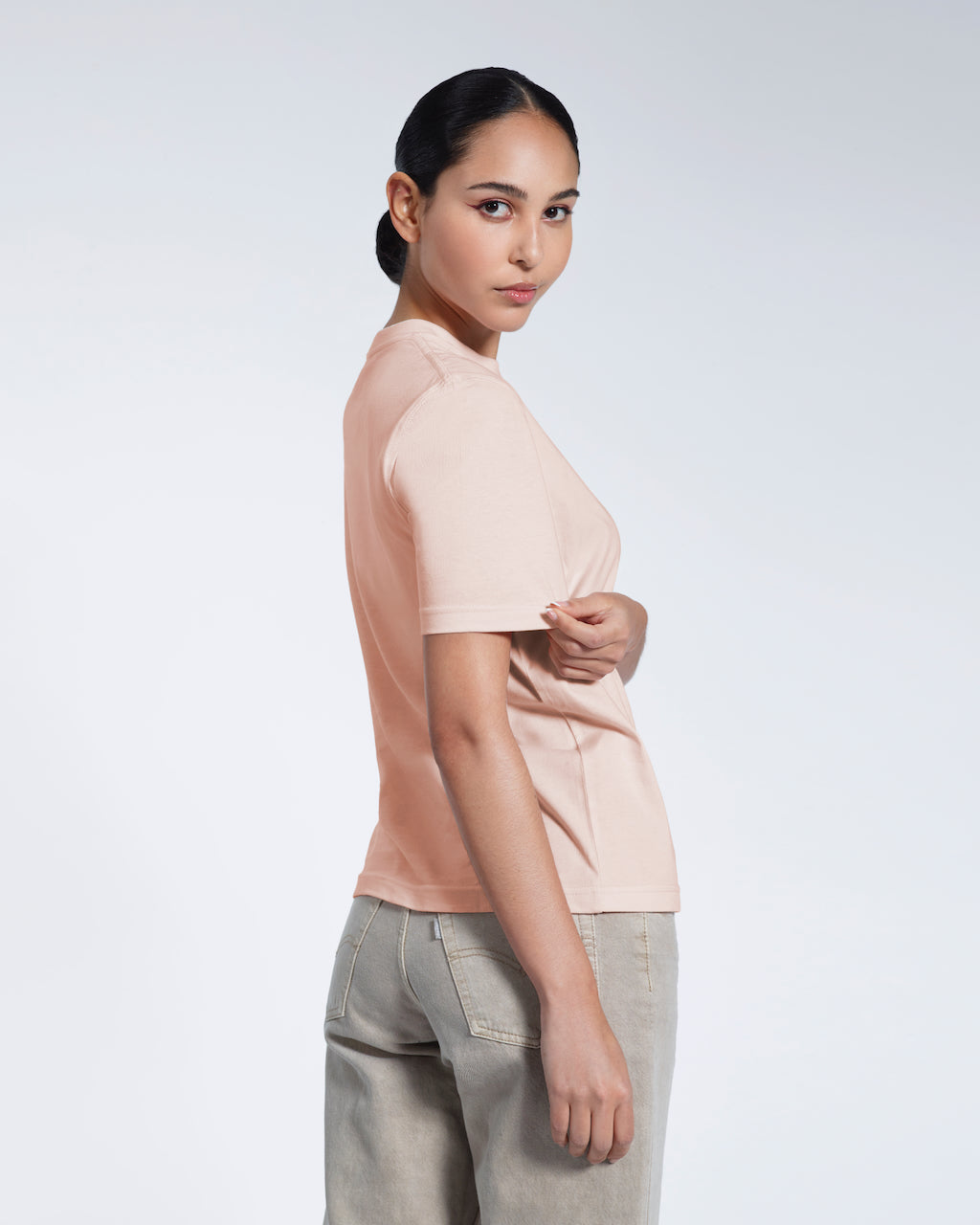 A woman turned towards the camera touching her sleeve, modelling a plain pink short sleeve organic cotton t shirt.