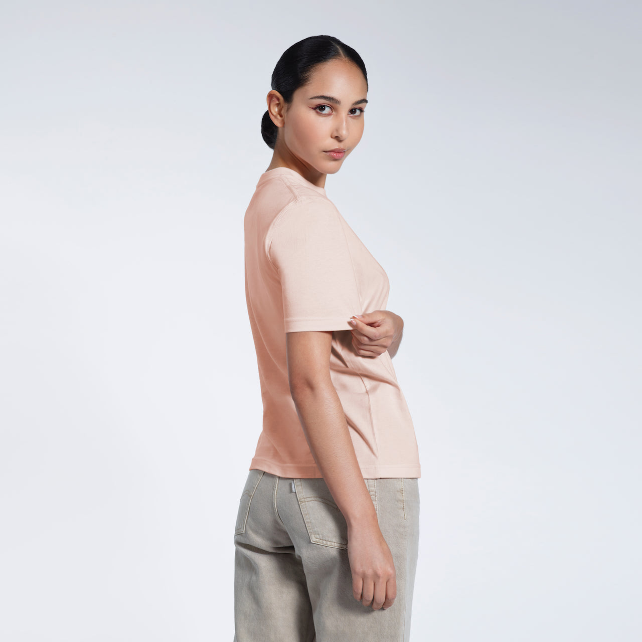 A woman turned towards the camera touching her sleeve, modelling a plain pink short sleeve organic cotton t shirt.