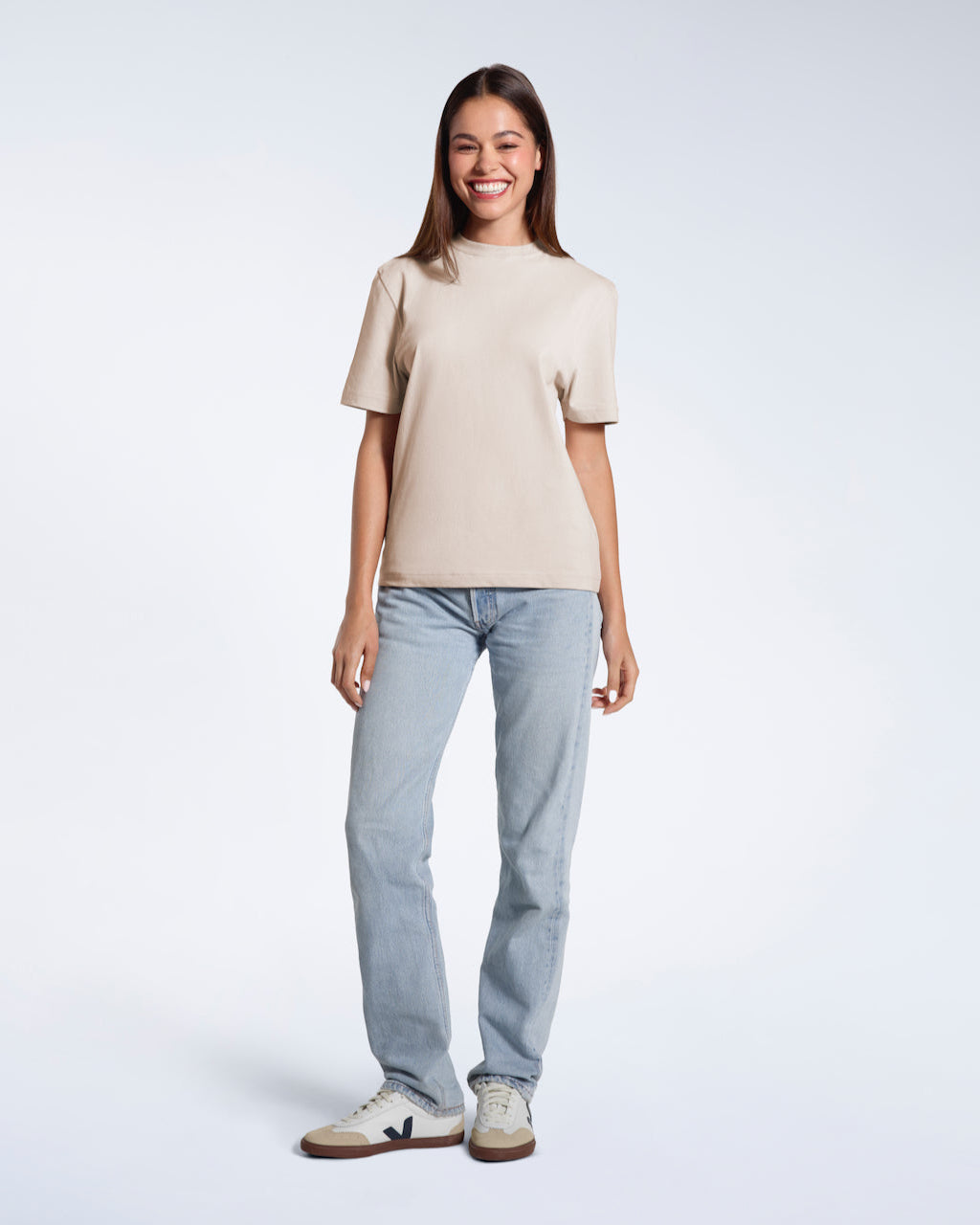 A full body photo of a woman smiling with her arms at her sides, modelling a plain stone coloured short sleeve organic cotton t shirt.