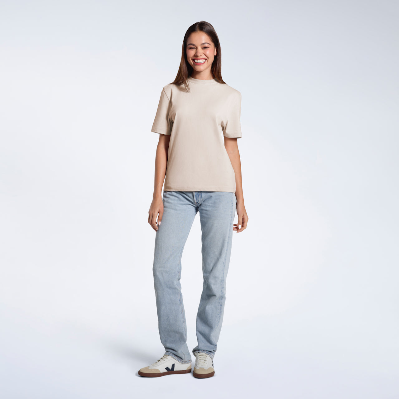 A full body photo of a woman smiling with her arms at her sides, modelling a plain stone coloured short sleeve organic cotton t shirt.