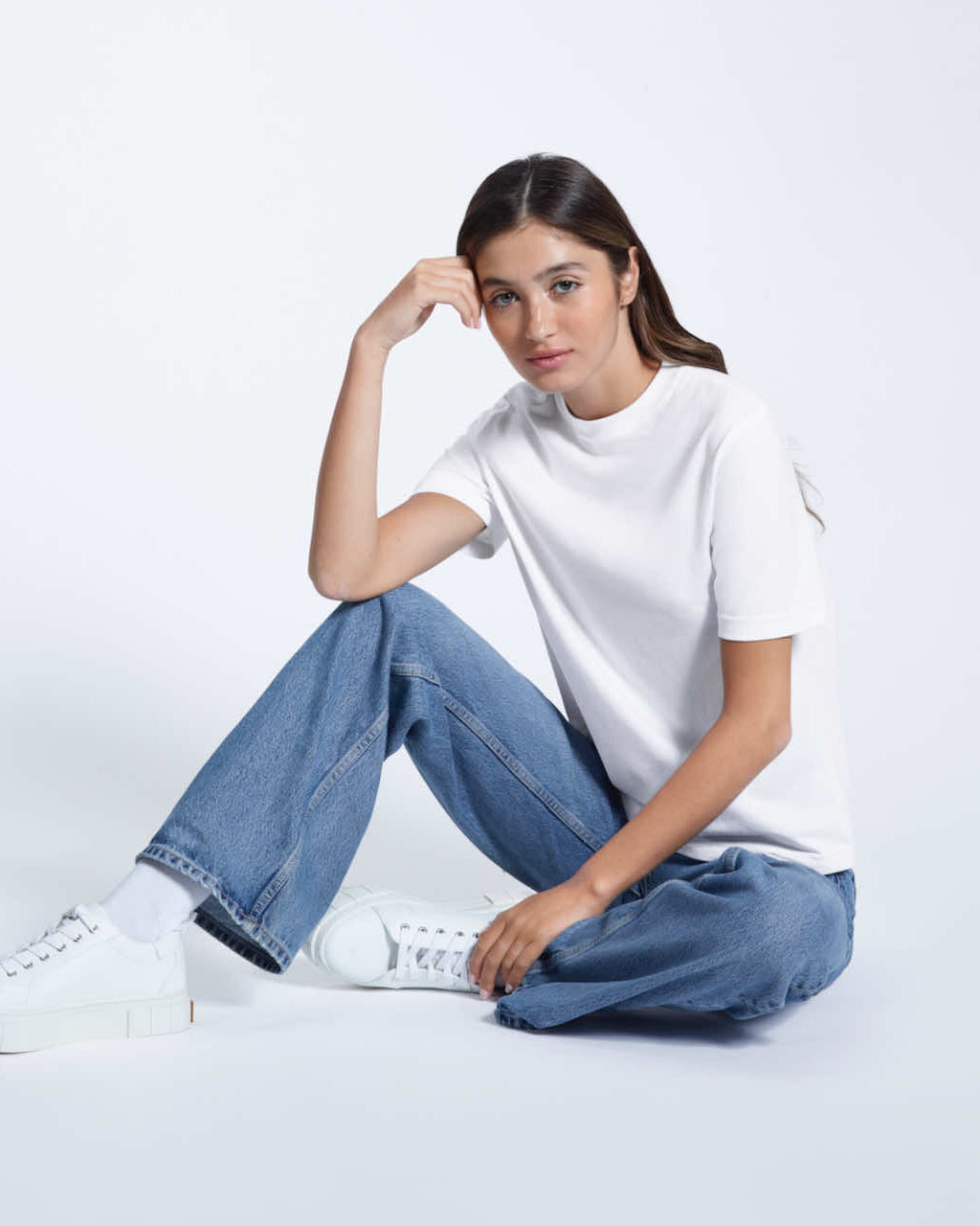 A woman sitting on the floor looking into the camera, modelling a plain white short sleeve organic cotton t shirt.