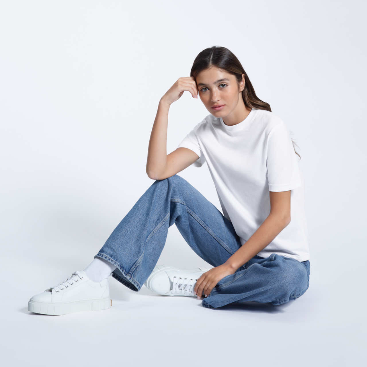 A woman sitting on the floor looking into the camera, modelling a plain white short sleeve organic cotton t shirt.