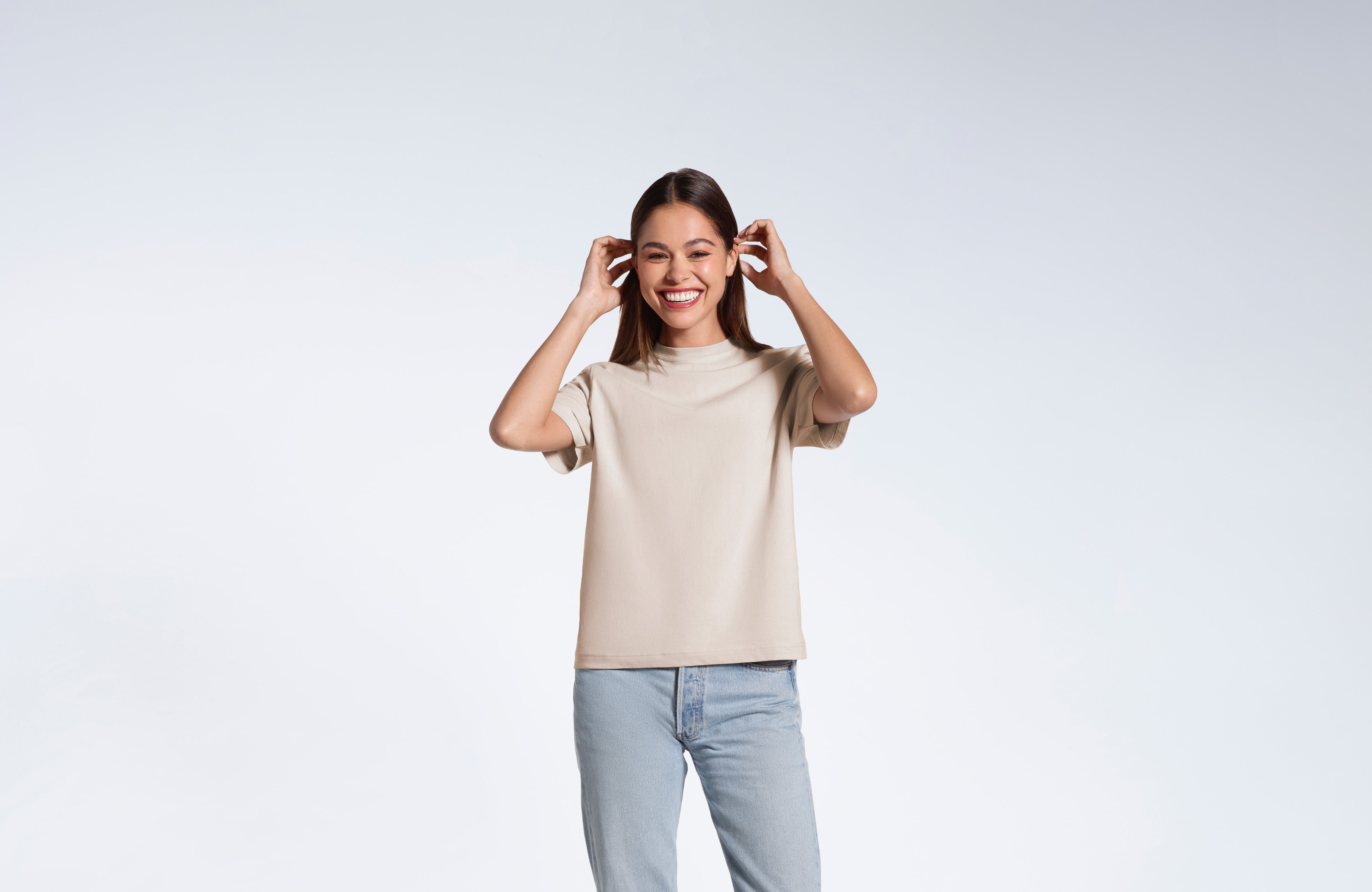 A woman touching her hair, smiling, modelling a stone coloured organic cotton, crew neck t shirt.