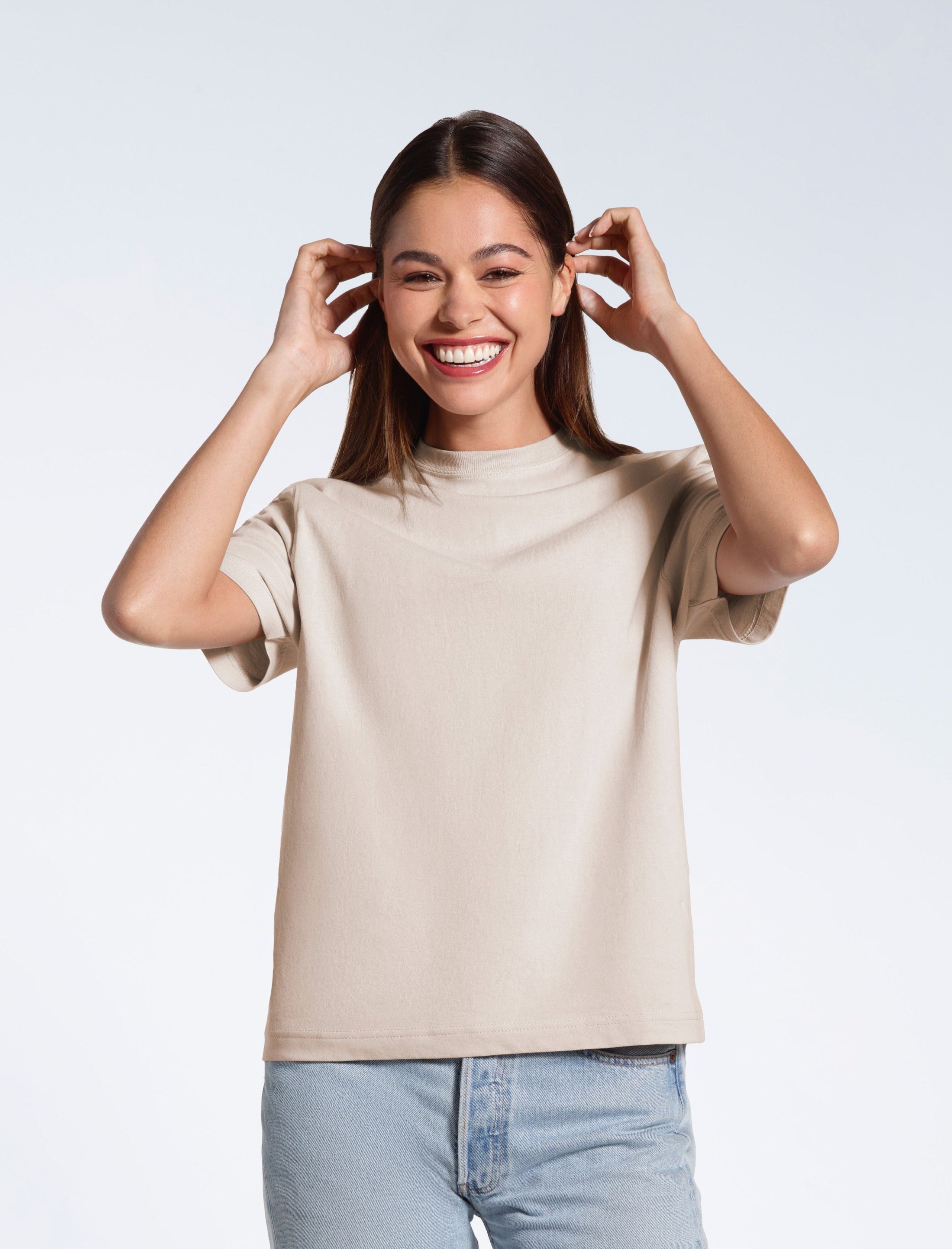 A woman touching her hair, smiling, modelling a stone coloured organic cotton, crew neck t shirt.