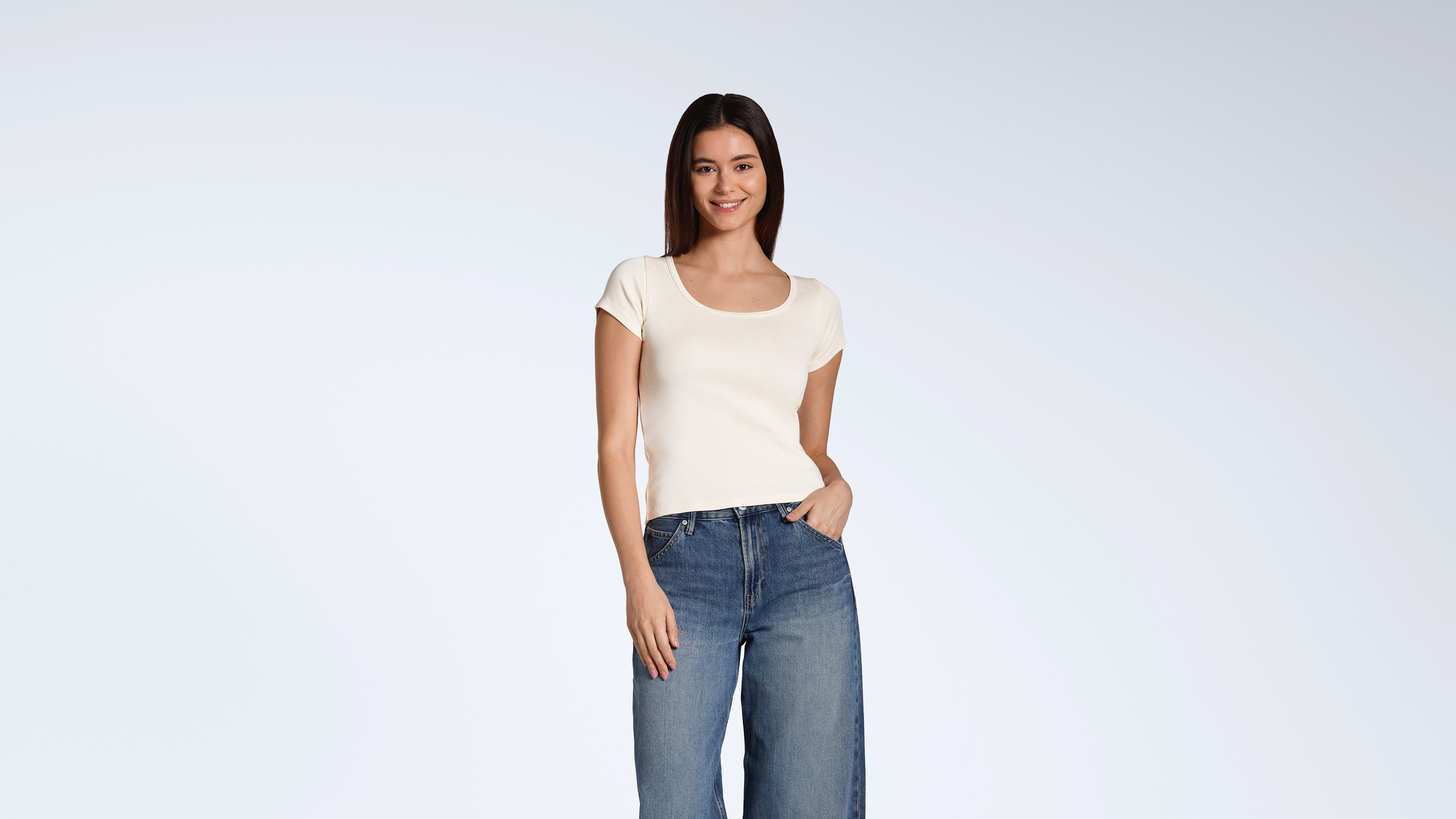A woman smiling into the camera with her hand in her pocket, modelling a plain, natural coloured, organic cotton, scoop neck t shirt.