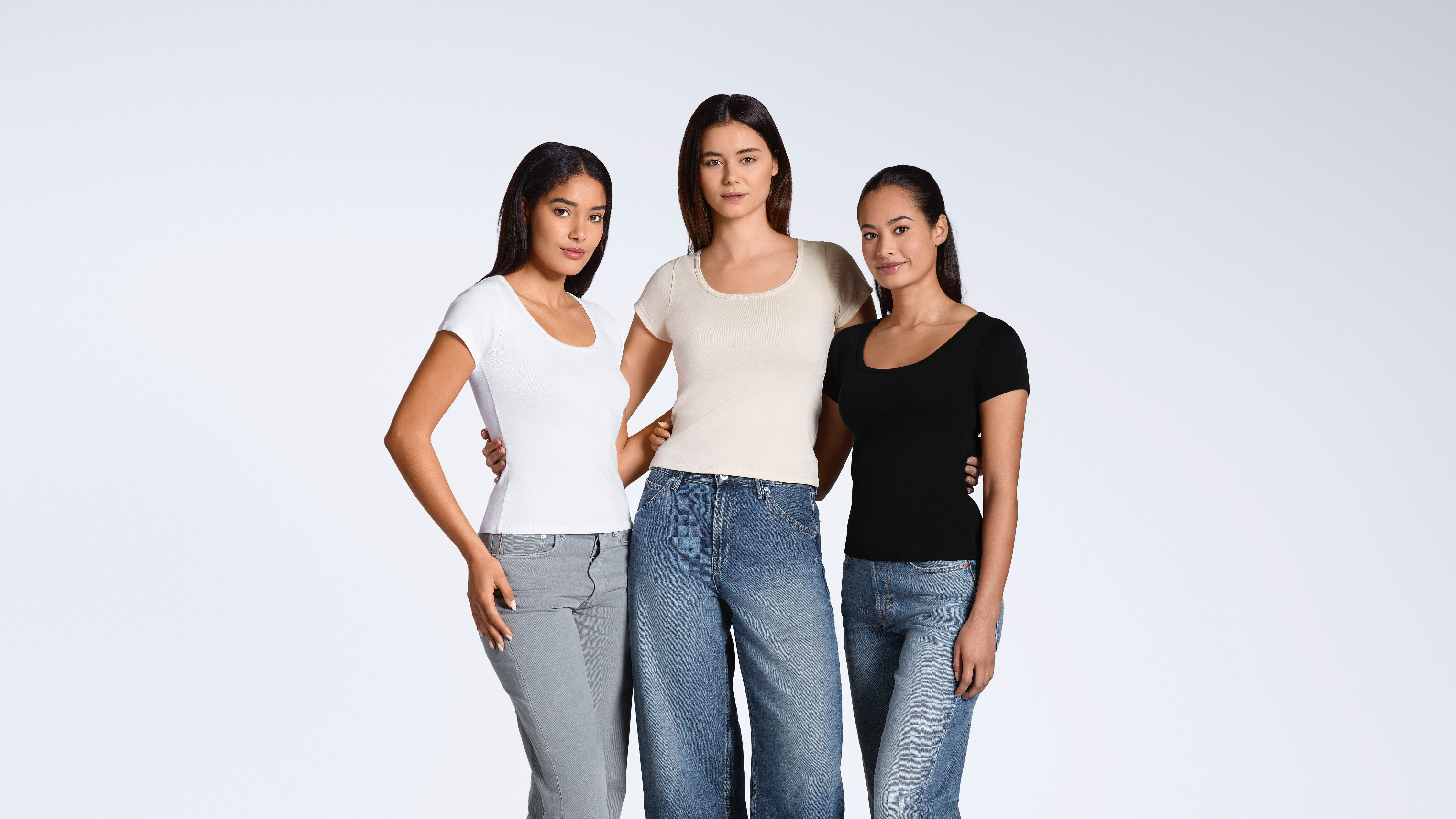 Three women with their arms round eachother looking into the camera, modelling plain white, natural and black, scoop neck, organic cotton t shirts.