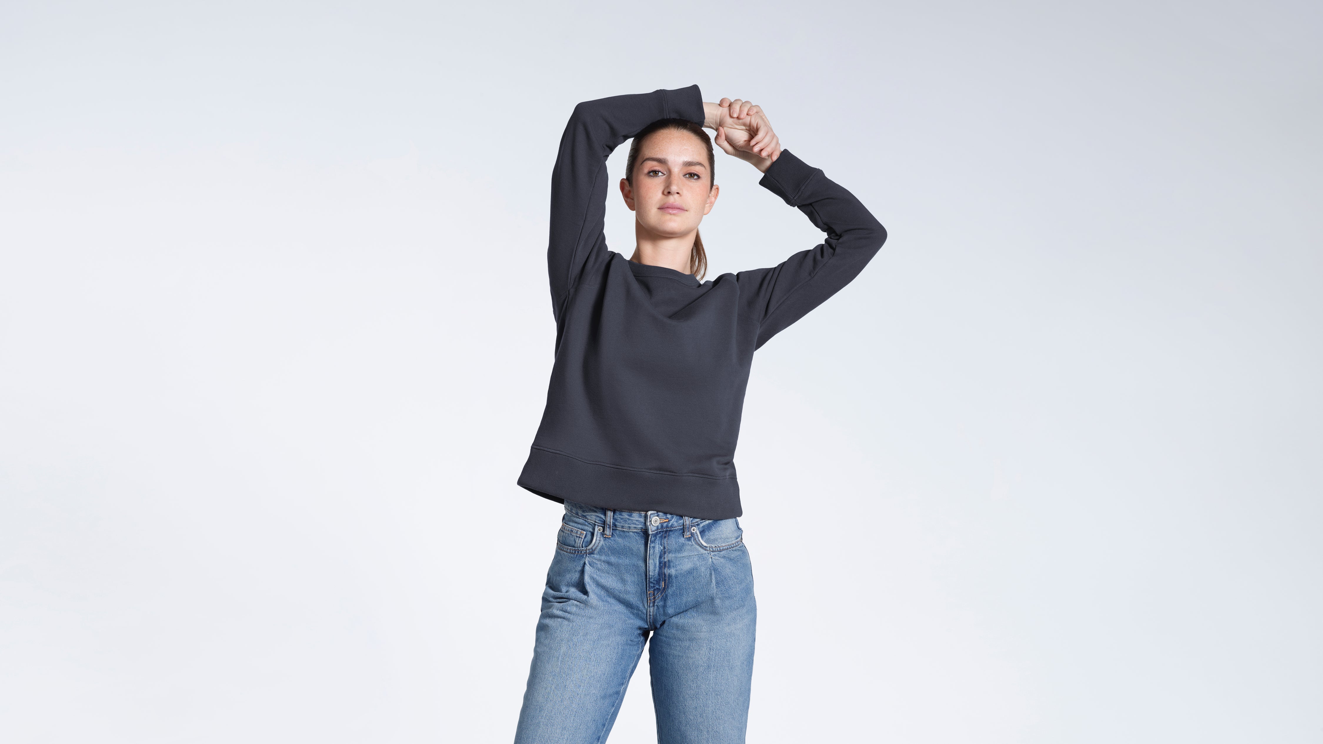 A woman looking into the camera with her hands above her head, modelling a plain dark grey french terry organic cotton sweatshirt.