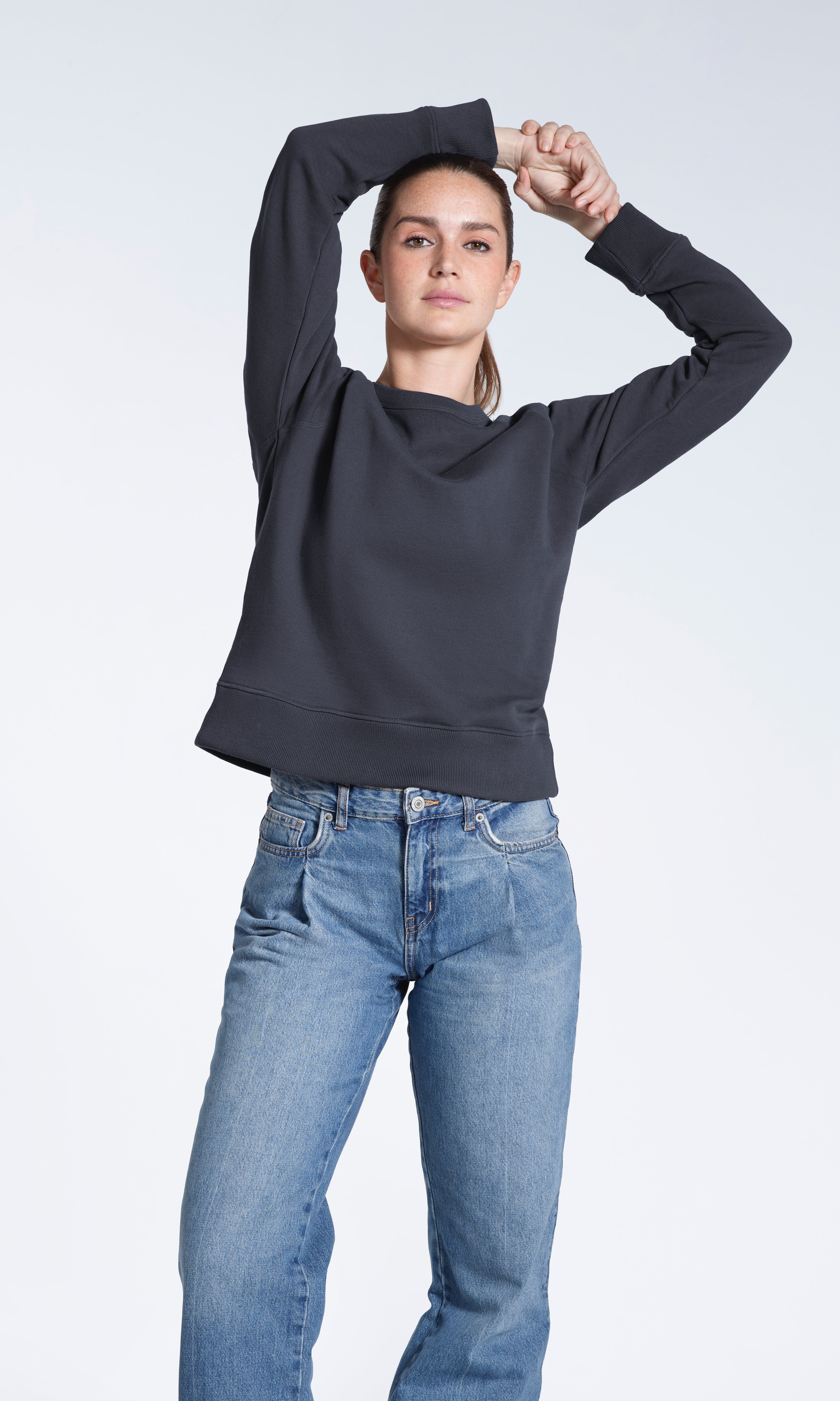 A woman looking into the camera with her hands above her head, modelling a plain dark grey french terry organic cotton sweatshirt.